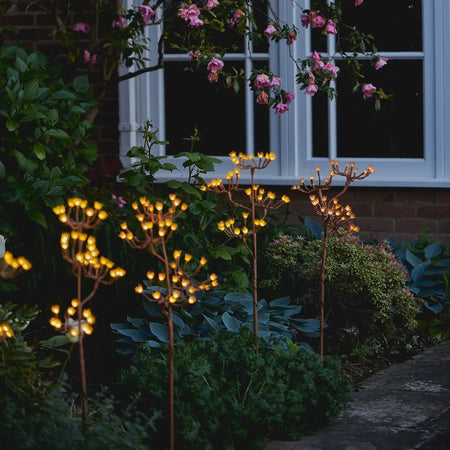 Charming Solar Lights styled to look like Fennell Branches lined up in a flower bed with their warm lights glowing in the dusk